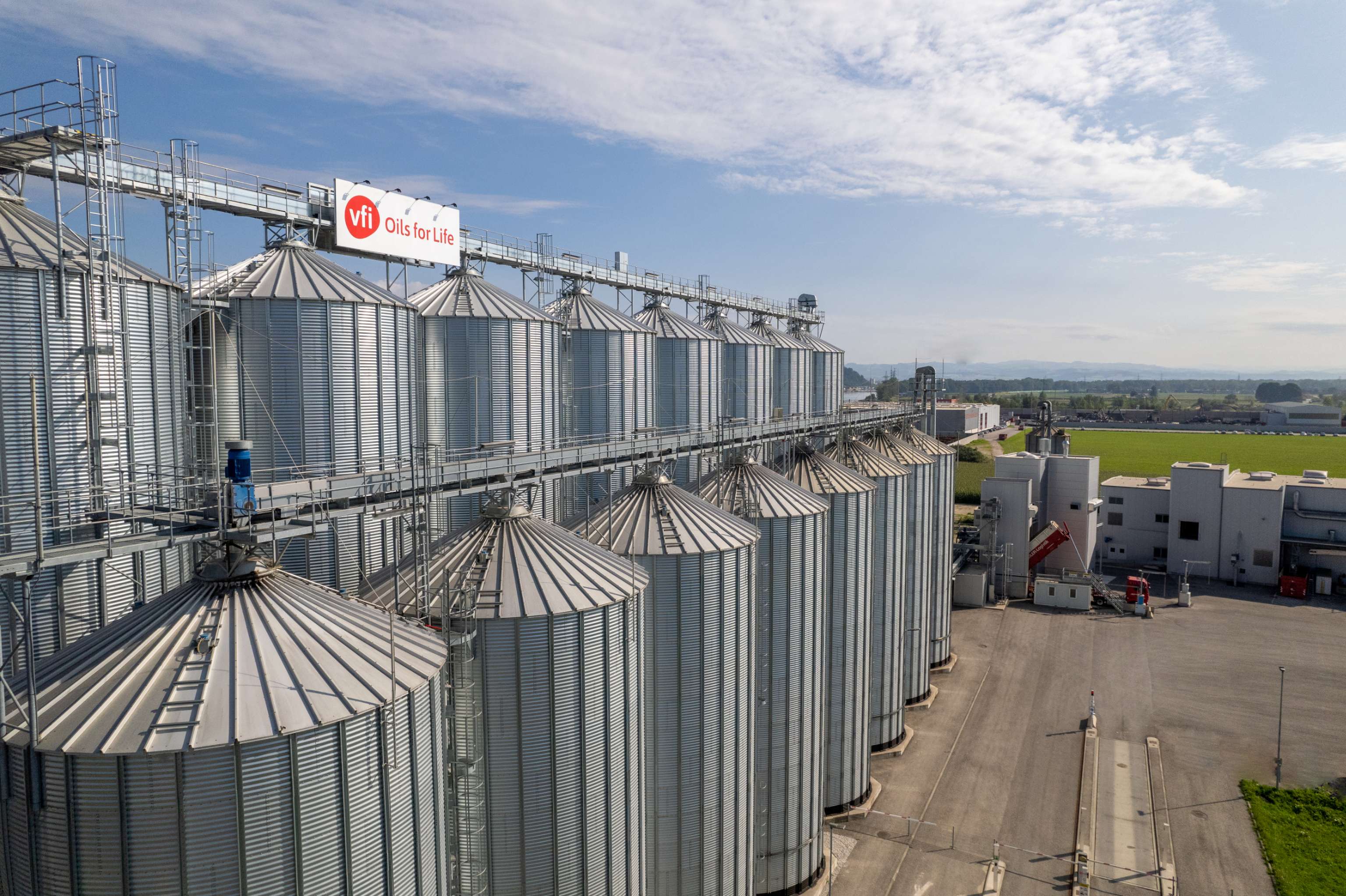 Aerial view of large metal silos at an agricultural facility with "Vfi Oils for Life" sign, under a partly cloudy sky.