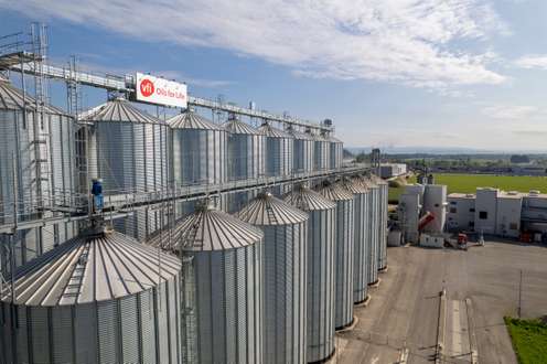 Aerial view of large metal silos at an agricultural facility with "Vfi Oils for Life" sign, under a partly cloudy sky.