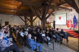 A speaker addresses an audience seated in a wooden-beamed hall, with flags and a podium on stage.