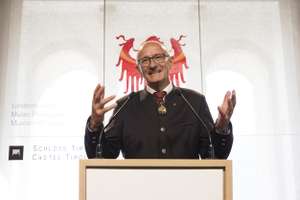 A man in a suit speaks at a podium with a coat of arms in the background.