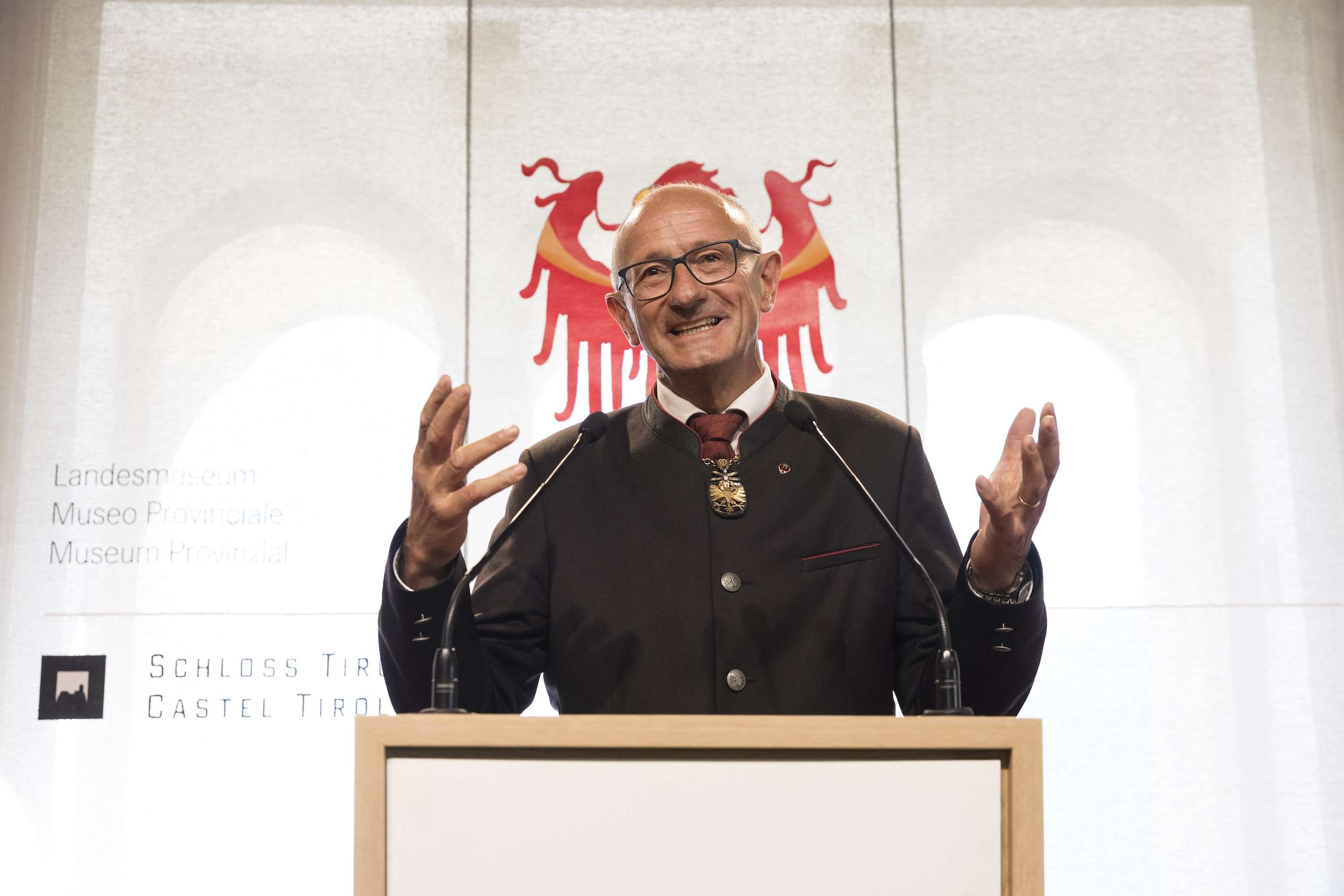 A man in a suit speaks at a podium with a coat of arms in the background.