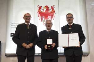 Three men in formal attire stand indoors; one holds a medal, another holds a certificate. A red eagle emblem is displayed in the background.