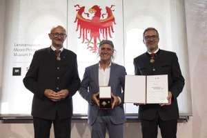 Three men in suits stand smiling; the man in the center holds a box with a medal. A certificate and emblem are displayed in the background.
