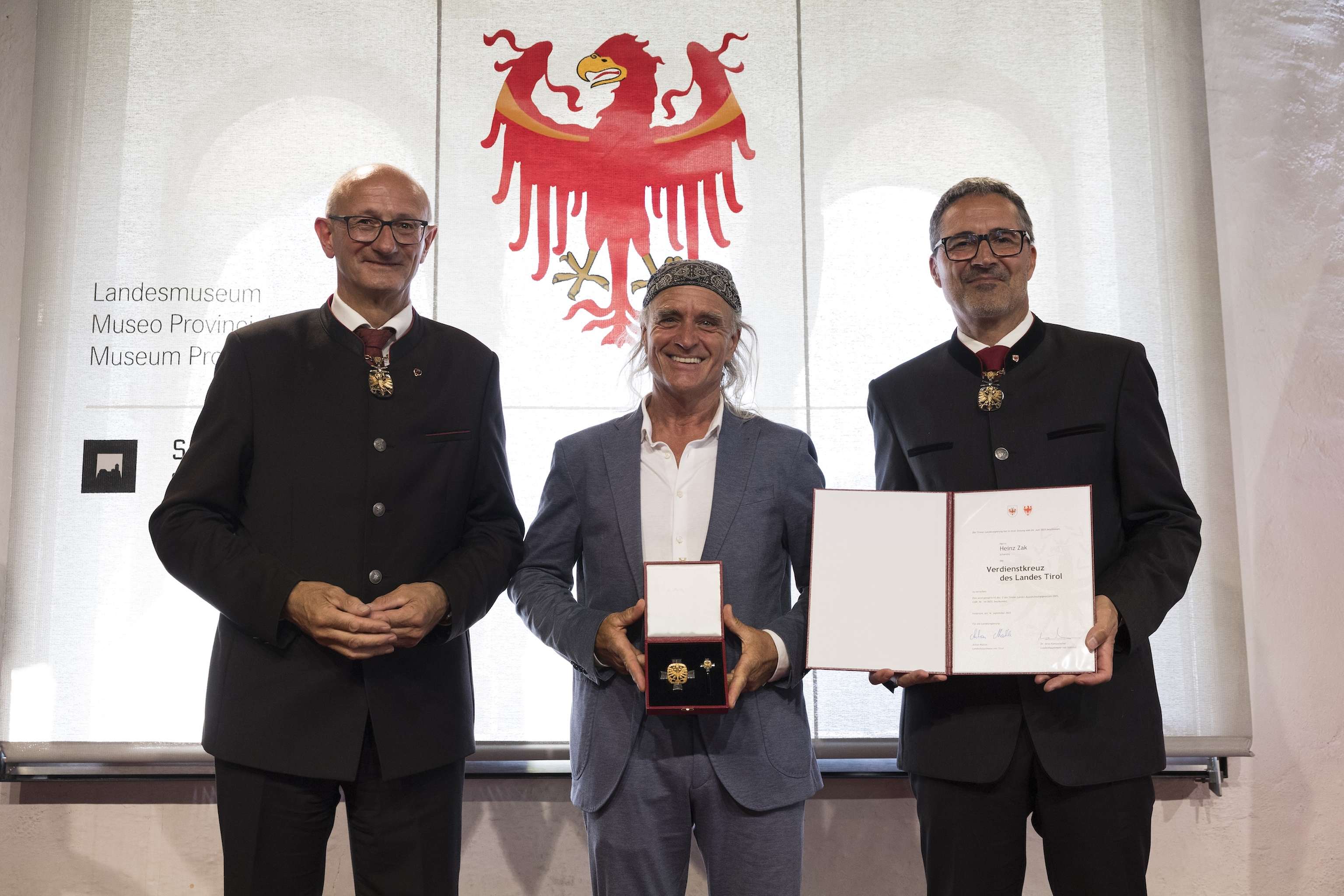 Three men in suits stand smiling; the man in the center holds a box with a medal. A certificate and emblem are displayed in the background.