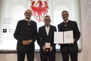 Three men in formal attire stand together; the center man holds a certificate and medal. An emblem with an eagle is displayed in the background.