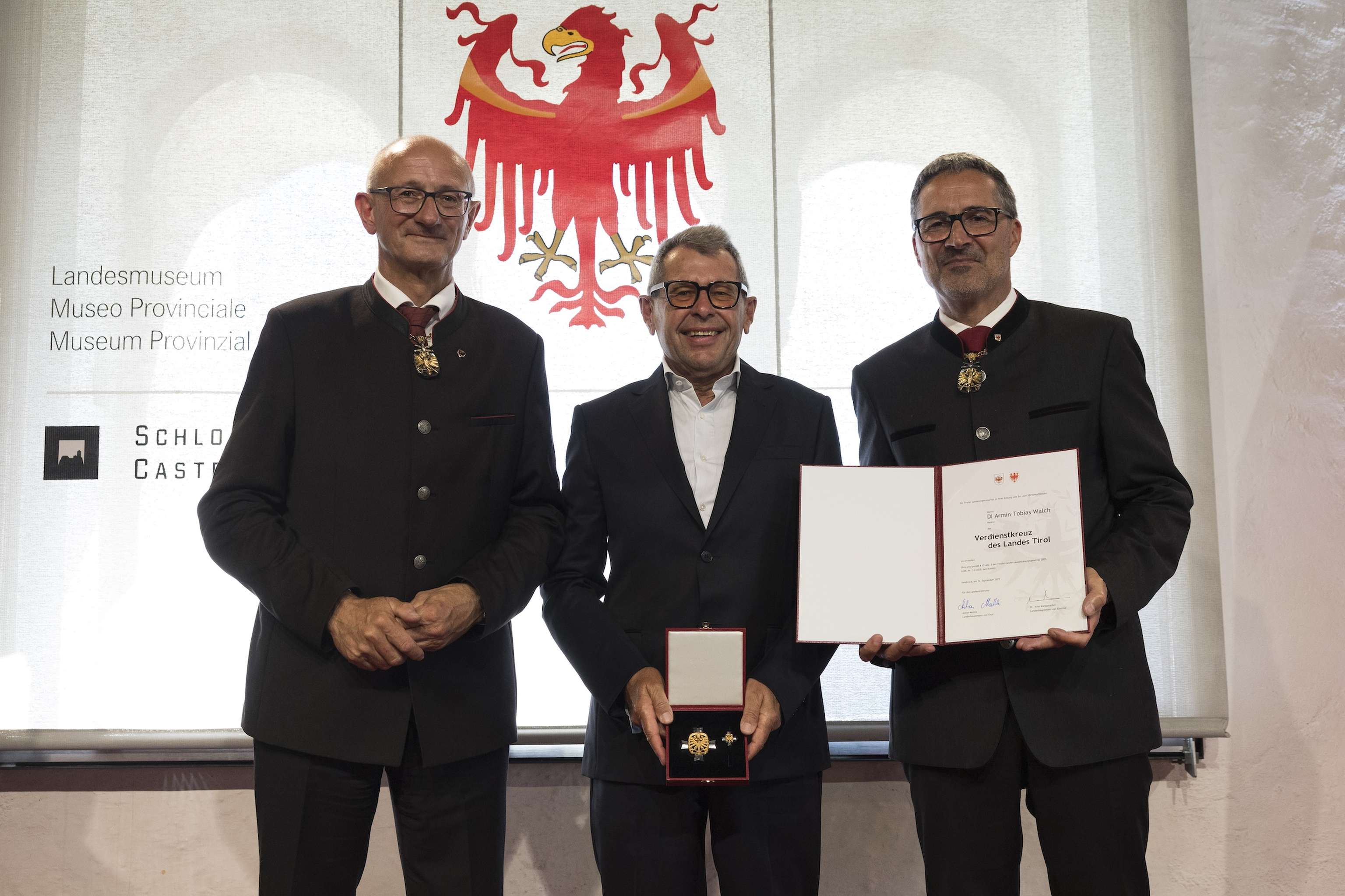Three men in formal attire stand together; the center man holds a certificate and medal. An emblem with an eagle is displayed in the background.