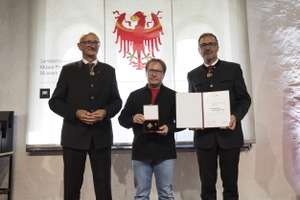 Three men stand together, one holding a medal and another holding a certificate, in front of a backdrop featuring a red eagle emblem.