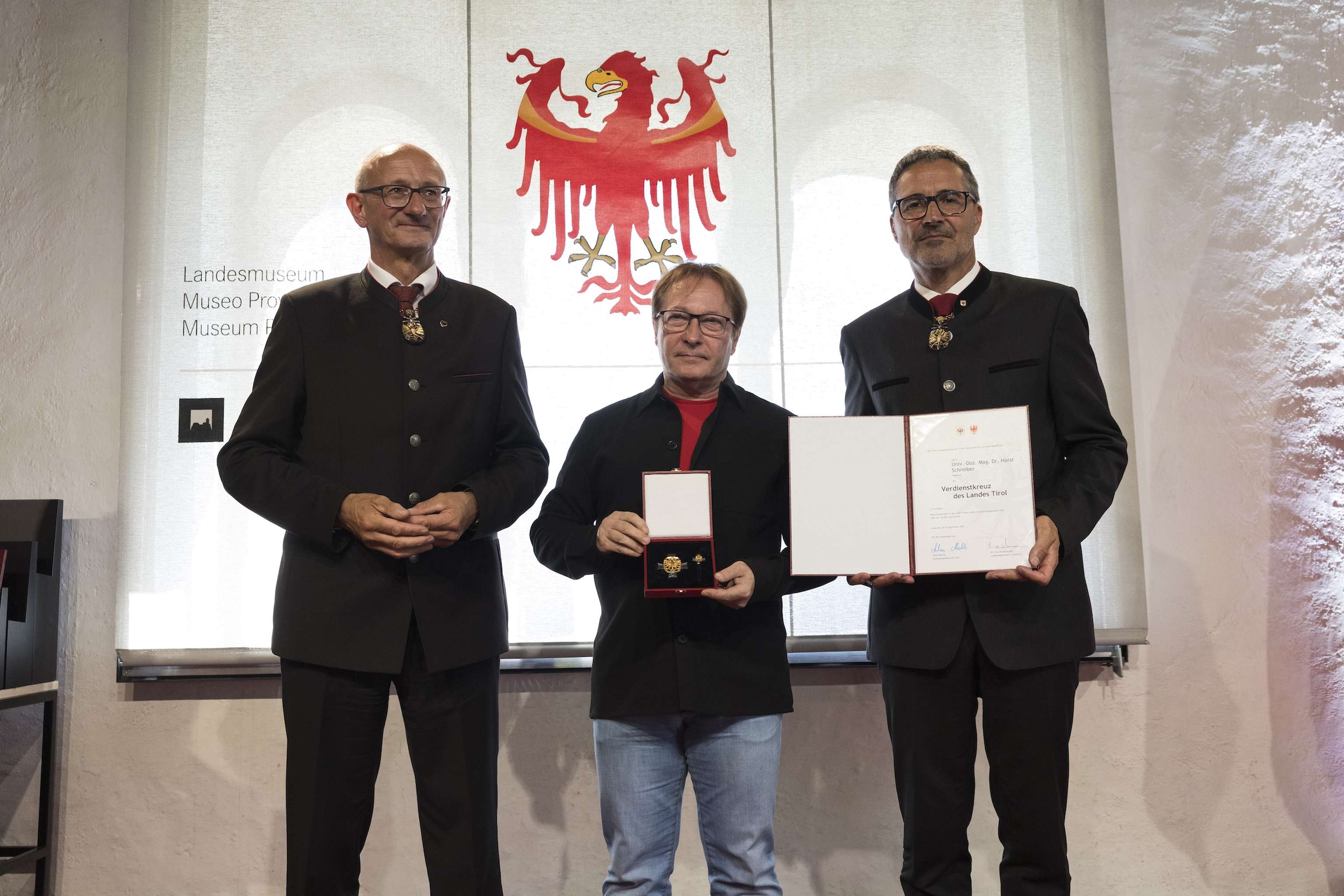 Three men stand together, one holding a medal and another holding a certificate, in front of a backdrop featuring a red eagle emblem.