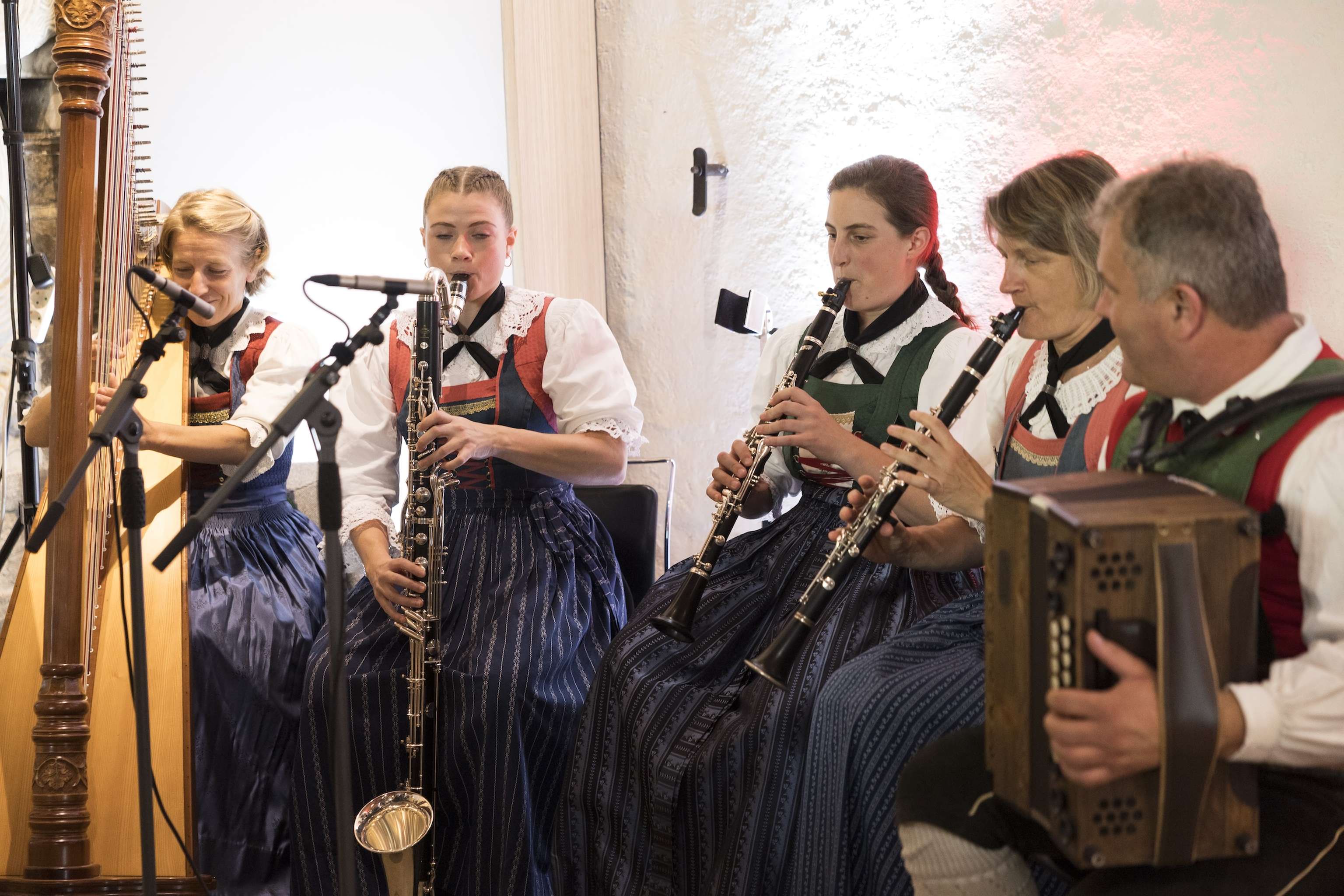 A group of musicians in traditional attire play various instruments, including clarinets, an accordion, and a harp, seated in a warmly lit room.