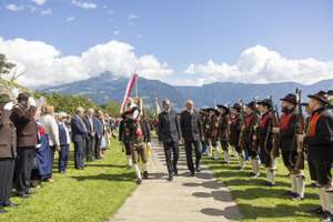 A formal procession with two men in suits walking between uniformed guards, set against a backdrop of mountains and a clear blue sky.