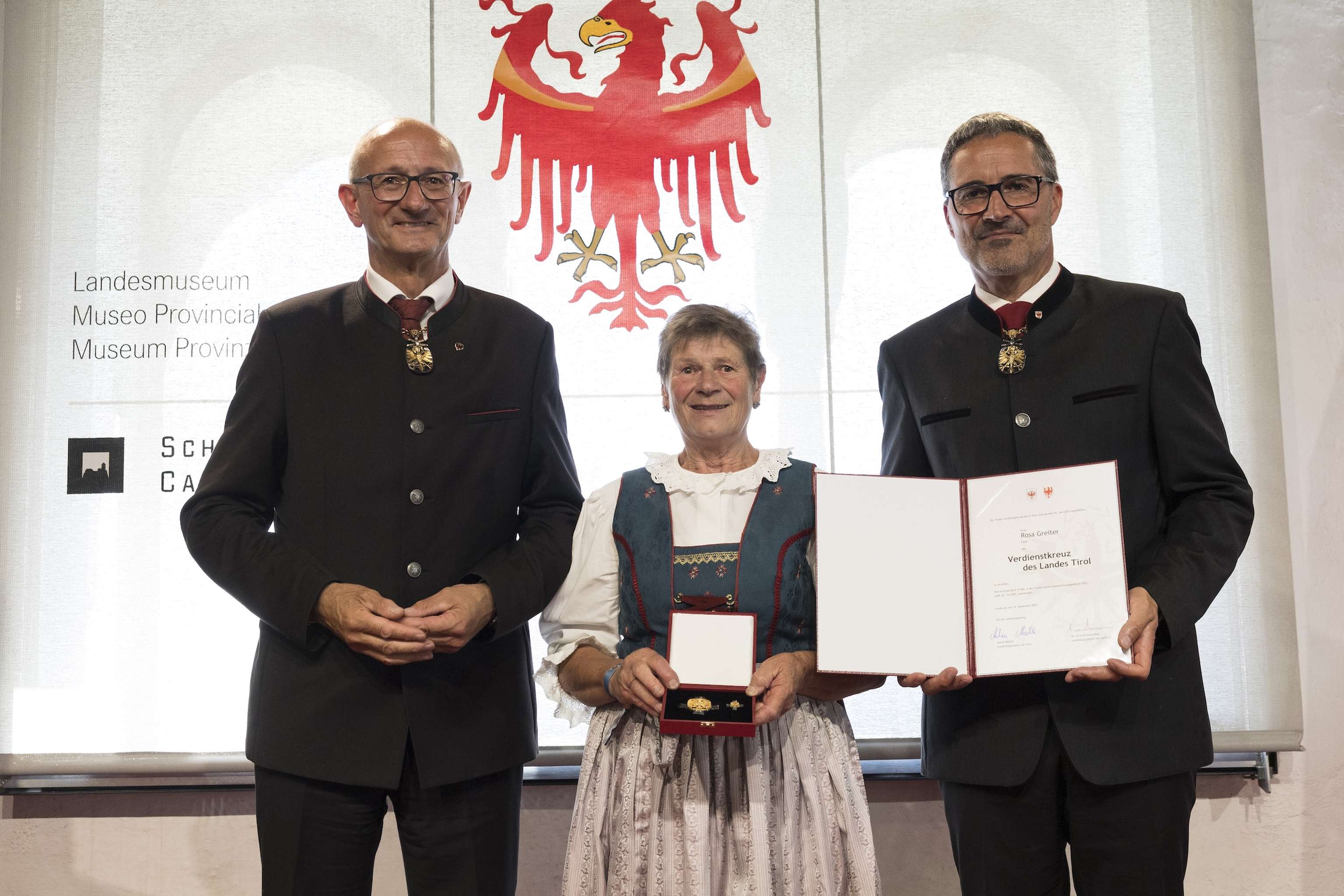 Three people in traditional attire stand indoors; one holds an open certificate and medal. A red eagle emblem is visible in the background.
