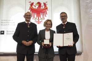 Three people stand smiling; the person in the center holds a medal, and the person on the right displays a certificate. A crest is visible in the background.
