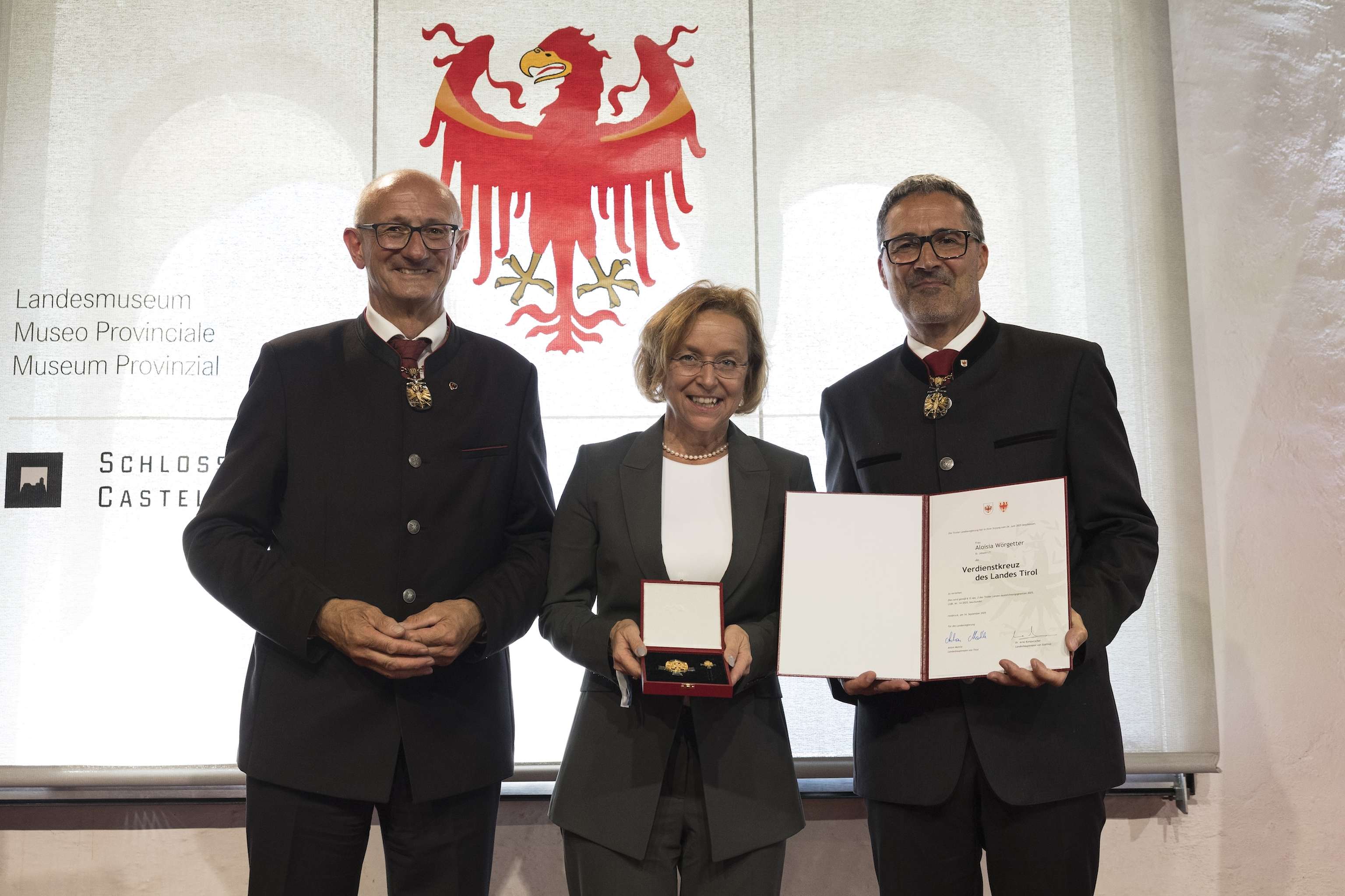 Three people stand smiling; the person in the center holds a medal, and the person on the right displays a certificate. A crest is visible in the background.