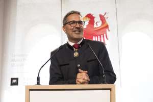 A man in a dark suit speaks at a podium with a red emblem behind him. He wears glasses and a decorative medallion.