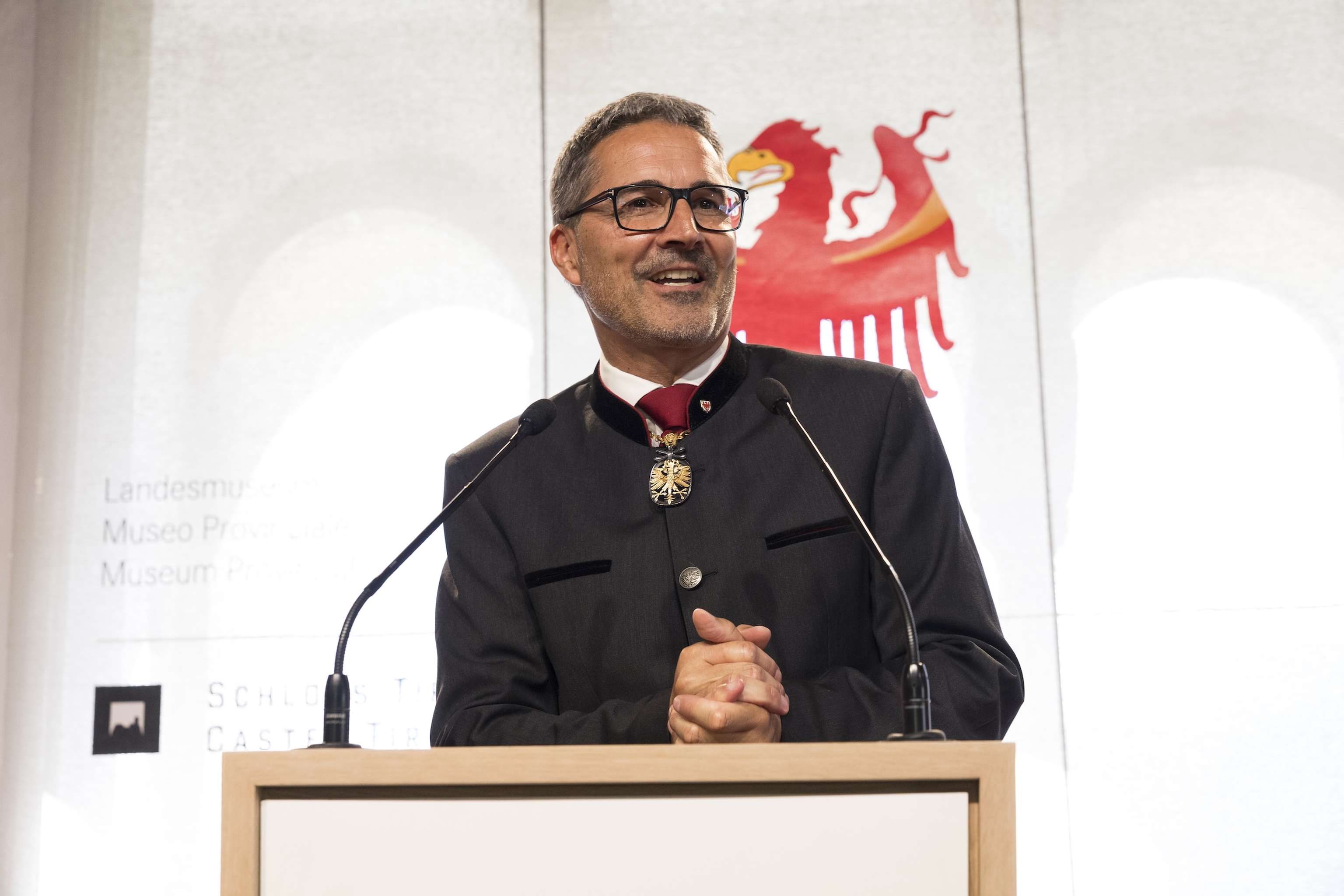 A man in a dark suit speaks at a podium with a red emblem behind him. He wears glasses and a decorative medallion.
