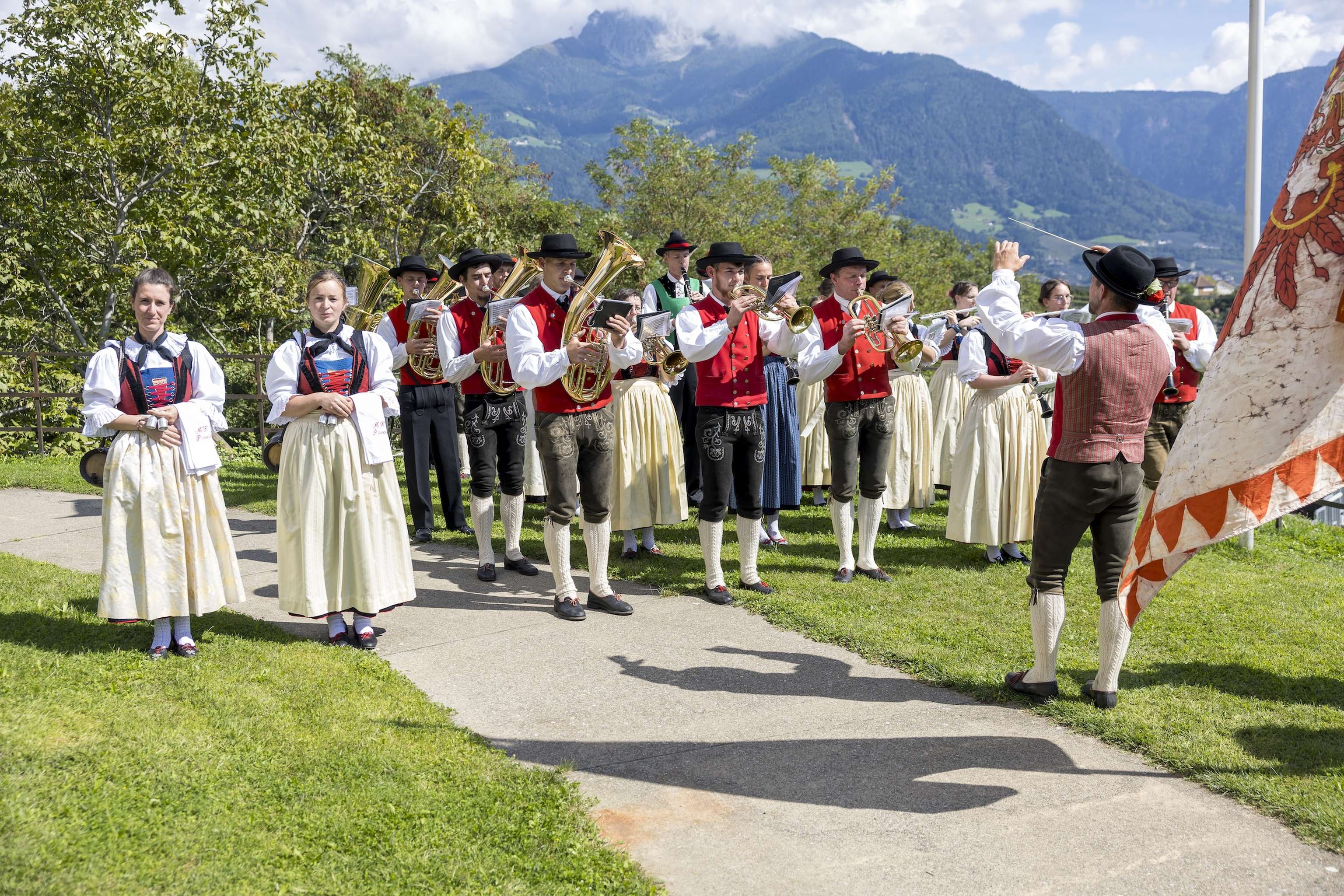 A traditional band in red vests and black hats plays outdoors, with mountains in the background. Women in traditional dresses stand nearby.