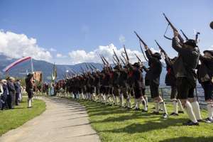 A line of people in traditional attire fire rifles in unison outdoors, with mountains and a blue sky in the background.