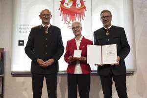 Three people standing, one holding a medal and certificate, in front of a backdrop featuring a red eagle emblem.
