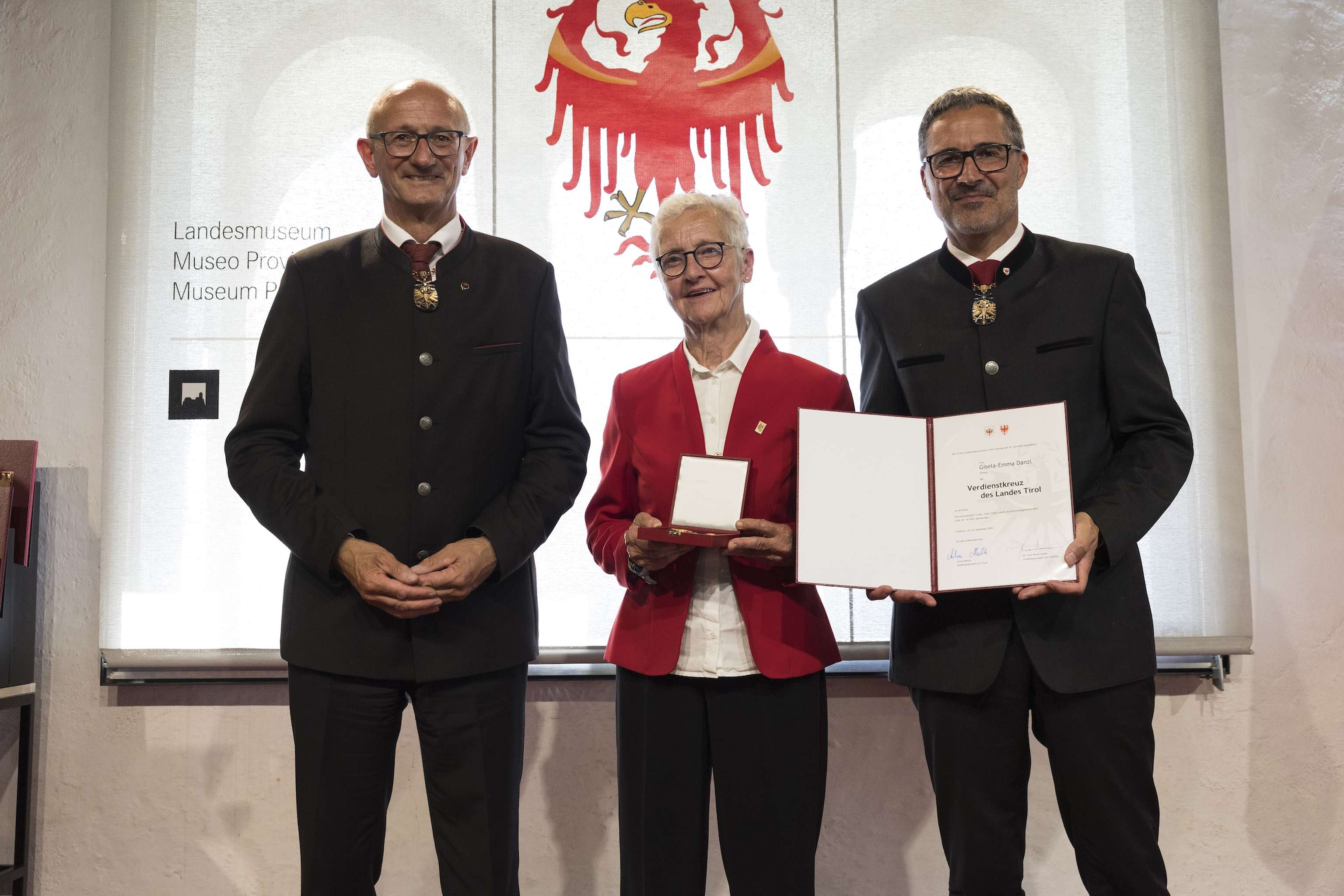 Three people standing, one holding a medal and certificate, in front of a backdrop featuring a red eagle emblem.