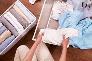 The Hands Of A Woman Sorting Through A Pile Of Clothes. There Are Piles Of Clothes Stacked In Metal Baskets On The Floor Nearby. The Concept Of Restoring Order, Cleaning, Cluttering, Organizing Space.