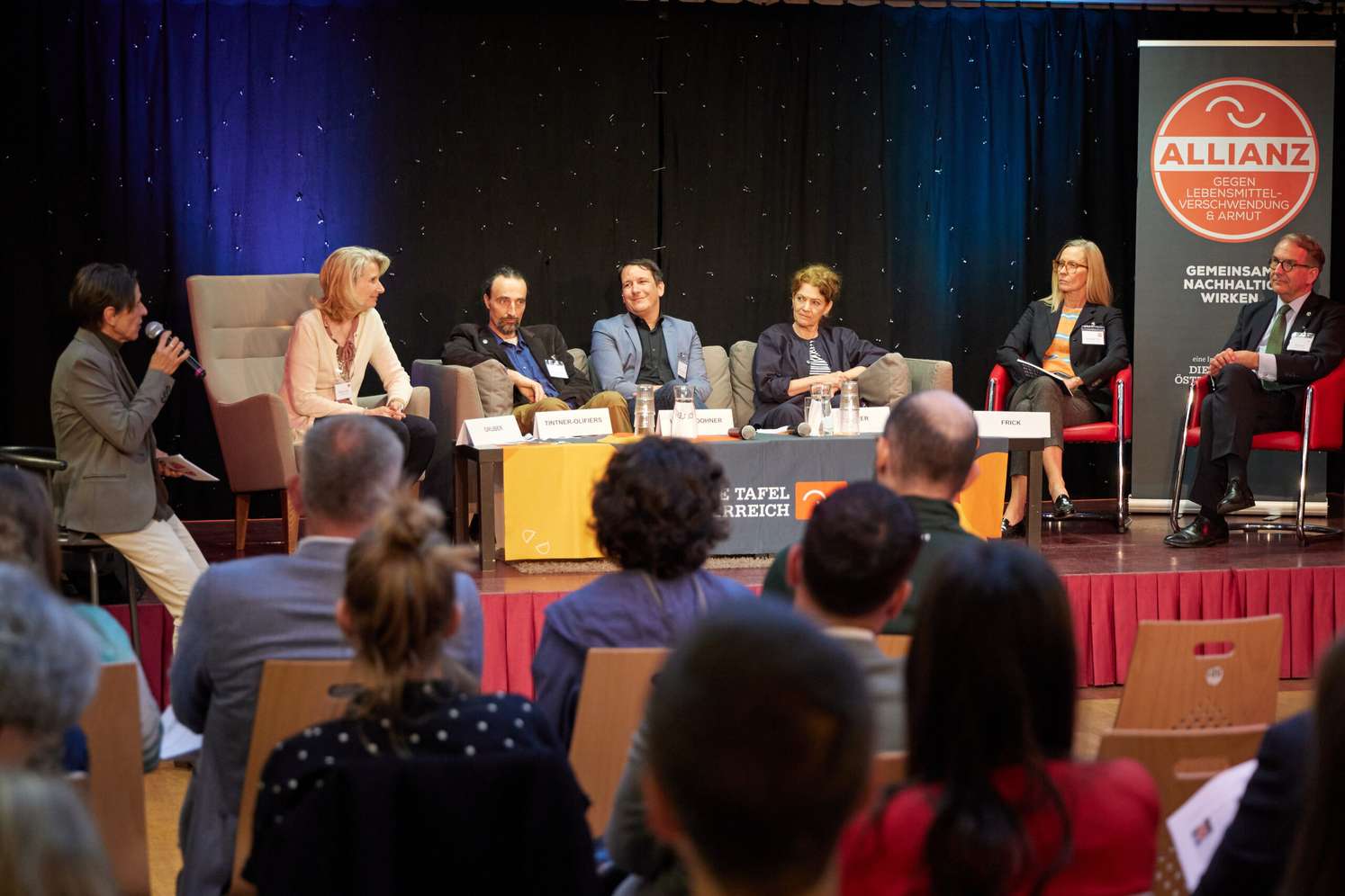 Podiumsdiskussion mit Personen. Ein Plakat rechts thematisiert die Allianz gegen Lebensmittelverschwendung und Armut. Publikum schaut zu.