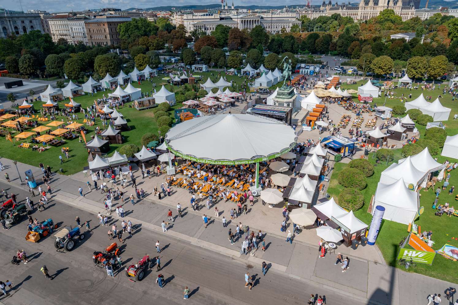 Luftaufnahme eines belebten Volksfestes in Wien mit weißen Zelten, Festzelt, Besuchern und historischen Gebäuden im Hintergrund.