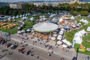 Luftaufnahme eines belebten Volksfestes in Wien mit weißen Zelten, Festzelt, Besuchern und historischen Gebäuden im Hintergrund.