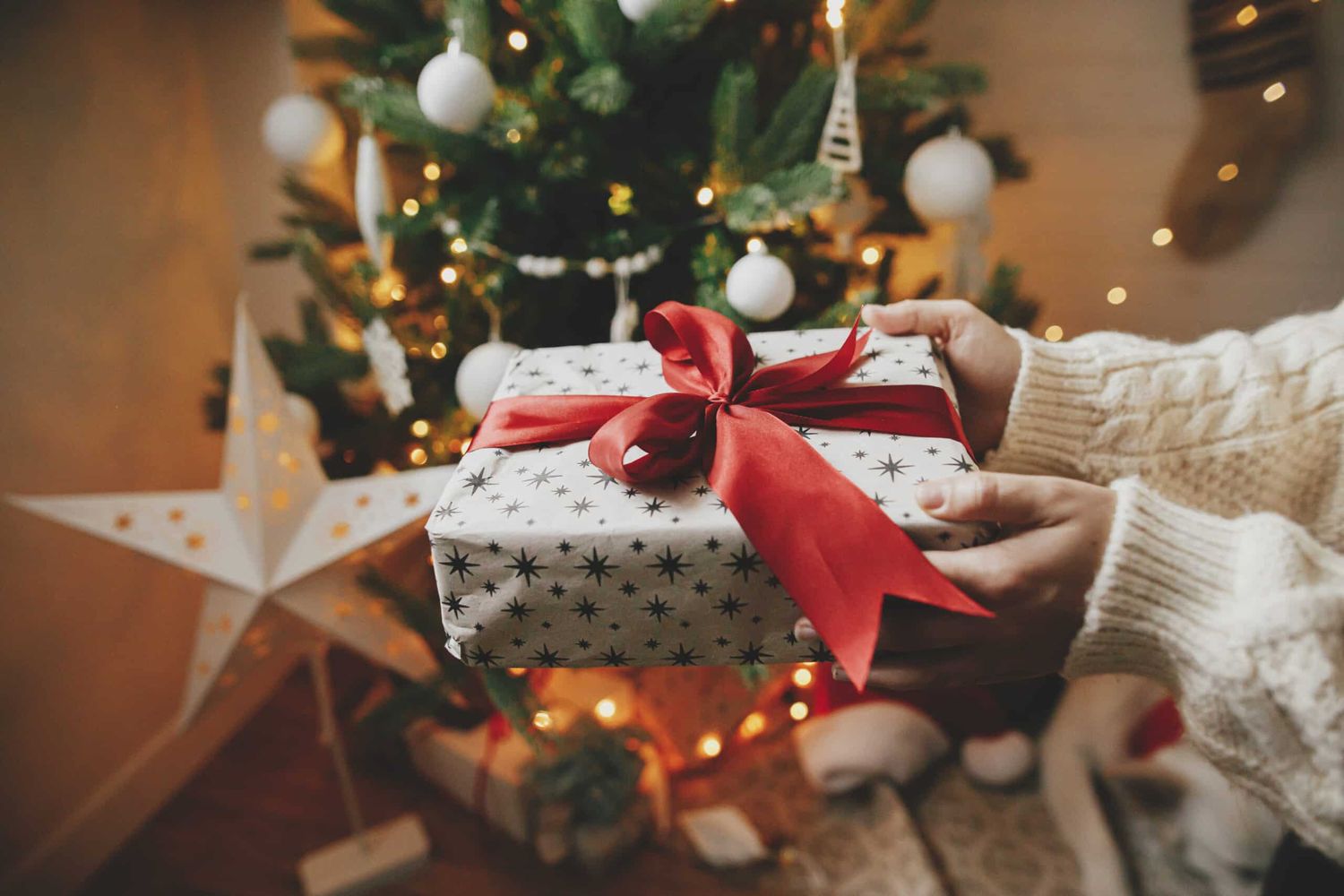 Wrapped Christmas Gift With Red Bow In Hands In Cozy Knitted Sweater On Background Of Christmas Tree With Lights. Stylish Woman Holding Present In Festive Decorated Room Close Up. Merry Christmas