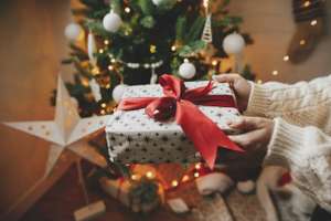 Wrapped Christmas Gift With Red Bow In Hands In Cozy Knitted Sweater On Background Of Christmas Tree With Lights. Stylish Woman Holding Present In Festive Decorated Room Close Up. Merry Christmas