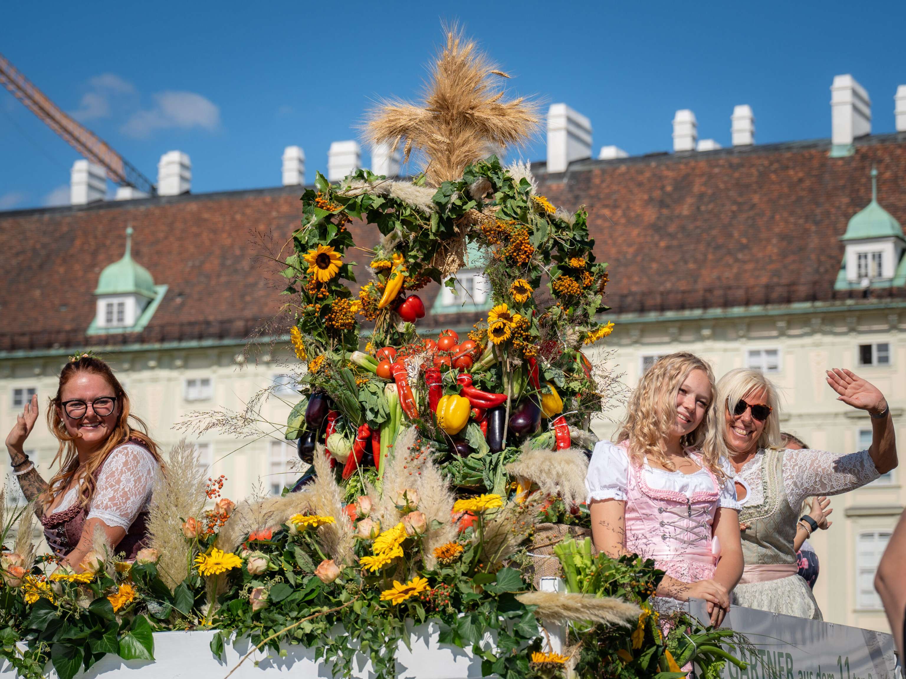 Frauen in Dirndl winken auf einem festlichen Erntedank-Festwagen mit Gemüse, Sonnenblumen und Getreide vor einem historischen Gebäude.
