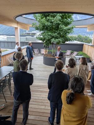 A group of people stand on a wooden deck under a covered area with a tree in the center, surrounded by mountains and buildings.