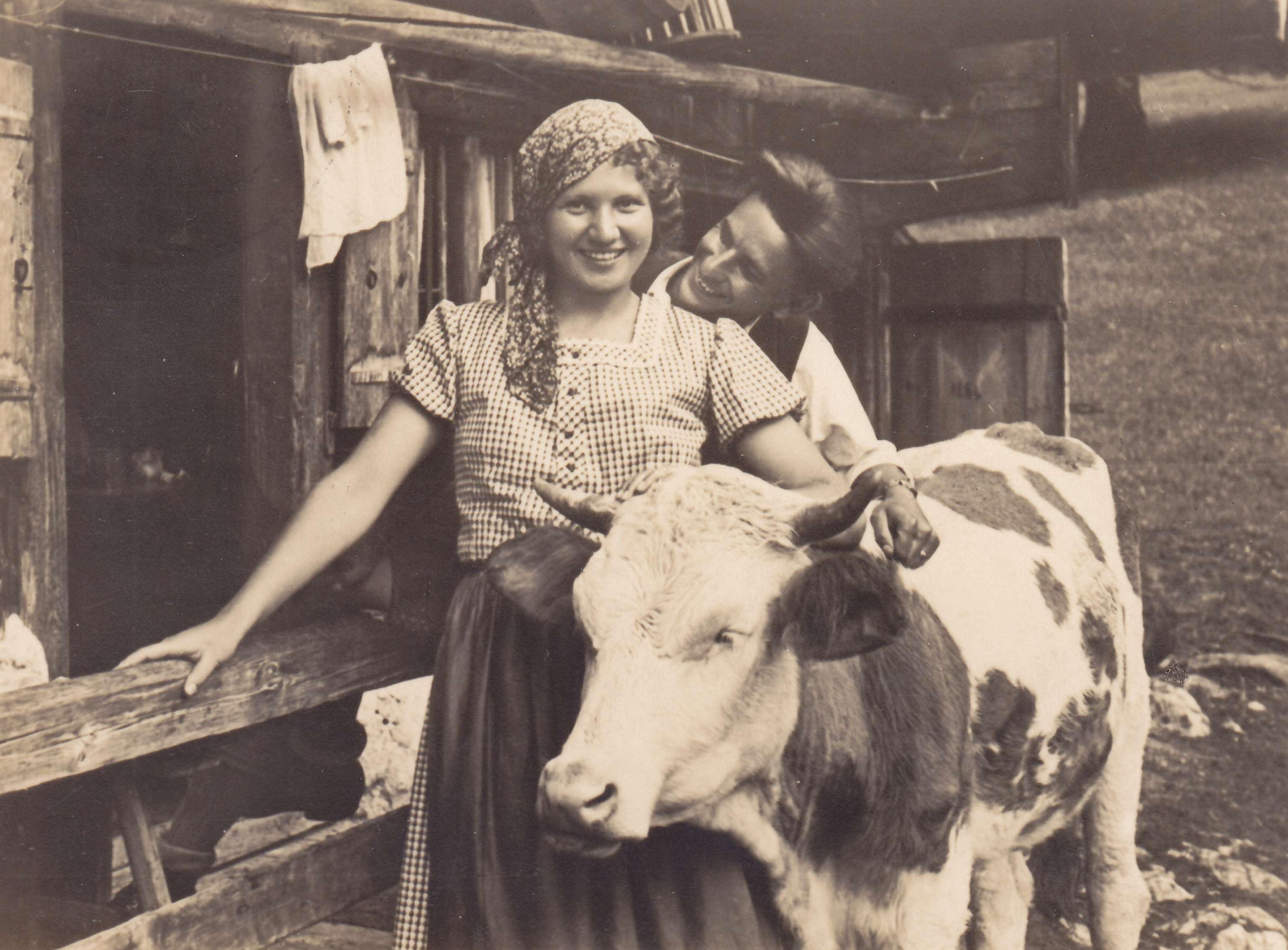Historisches Schwarzweißfoto zweier lächelnder Frauen in Bauerntracht neben einer Kuh vor einem Holzstall.
