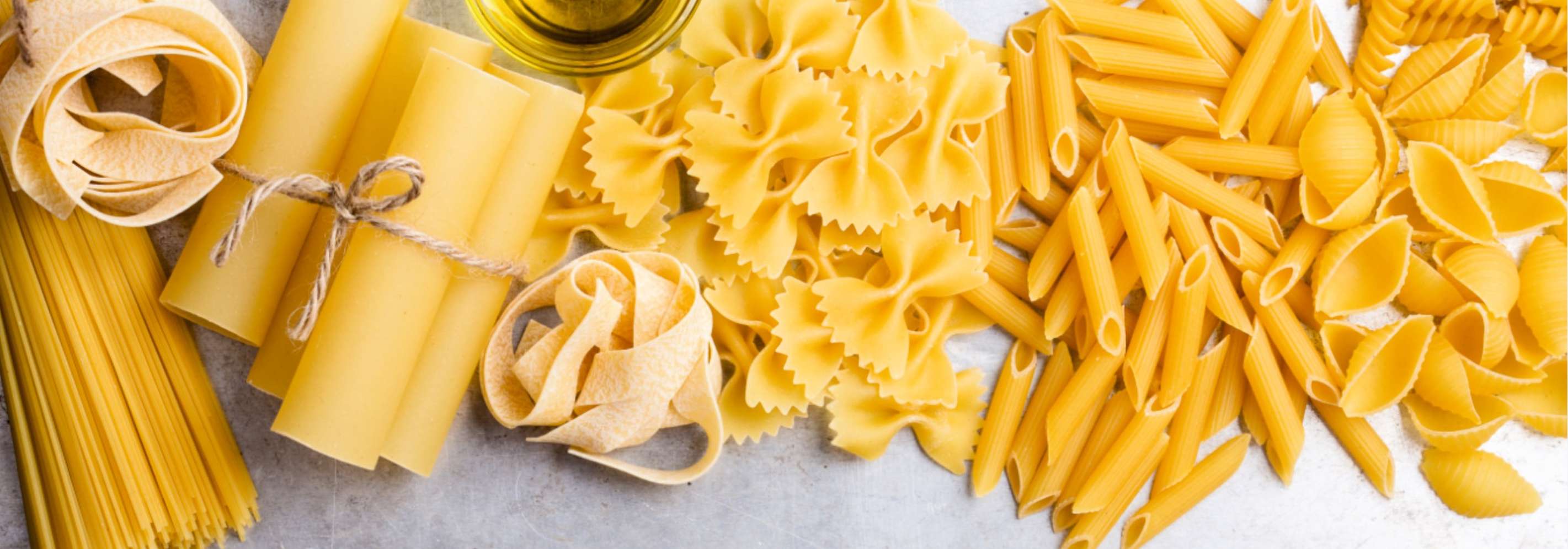 Verschiedene ungekochte Nudelsorten: Spaghetti, Lasagne, Farfalle, Penne und Muschelnudeln auf hellem Untergrund mit Olivenöl.