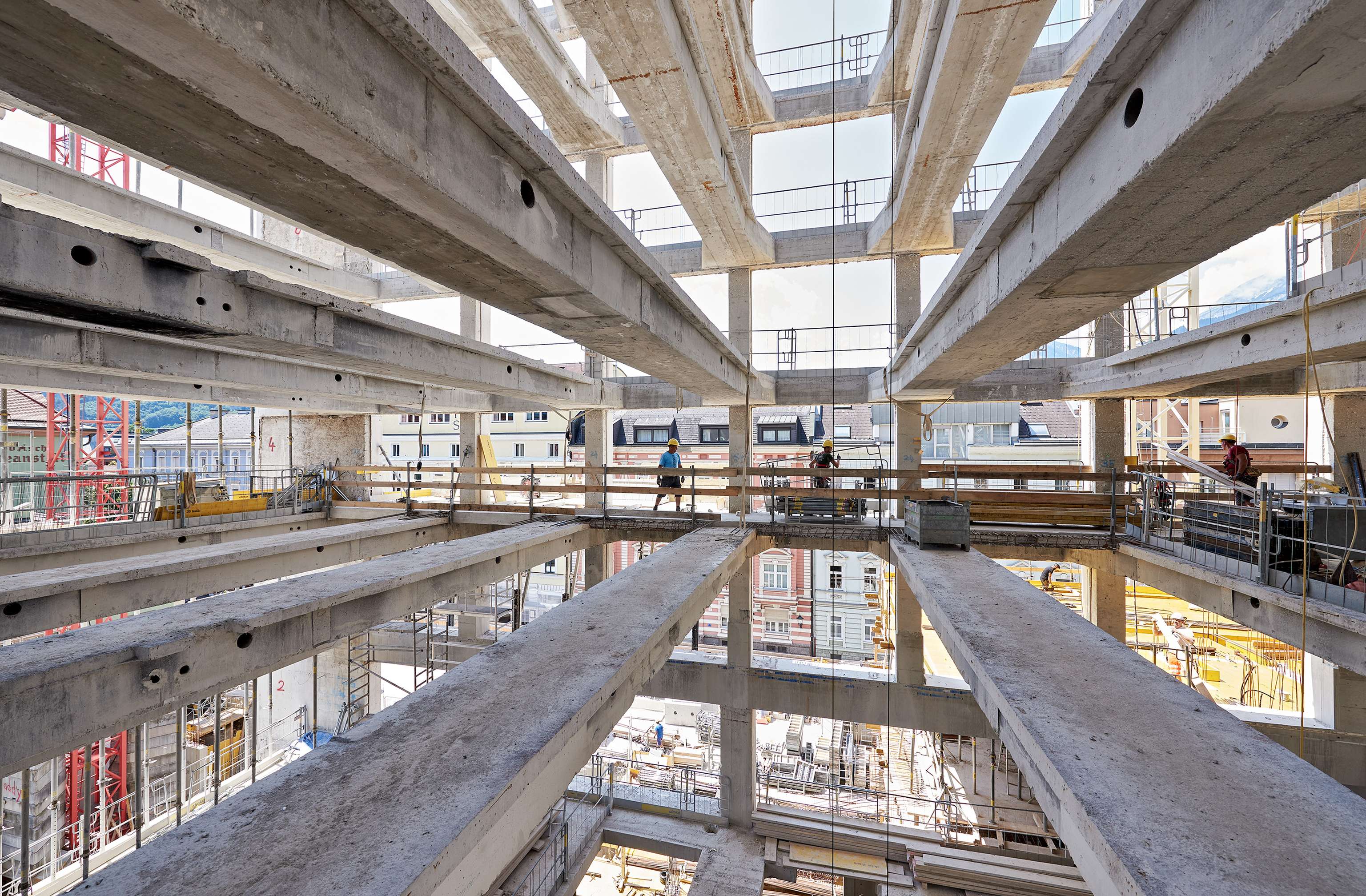 Construction site with multiple concrete floors and scaffolding, workers visible, and cranes in the background under a clear sky.