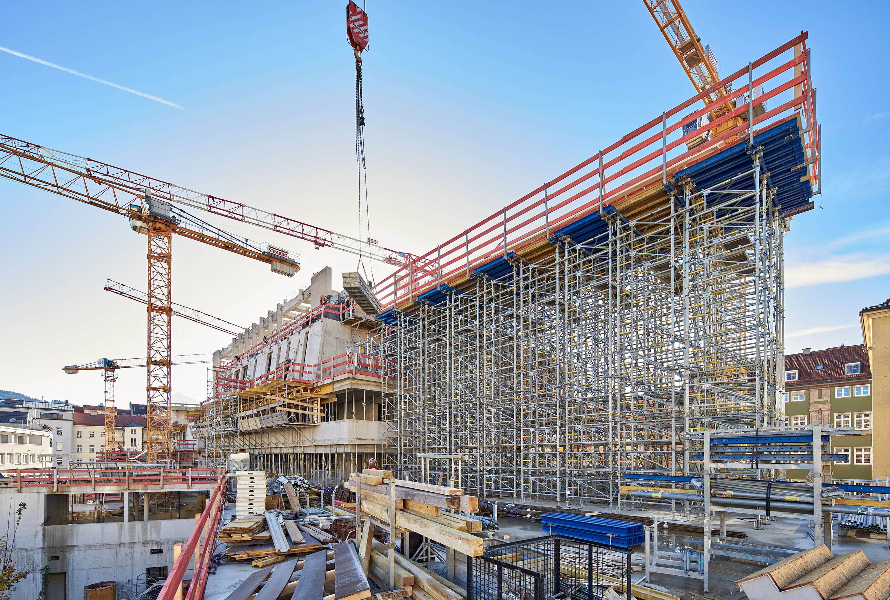 Construction site with multiple cranes, scaffolding, and building materials. Partially built structure under a clear blue sky.