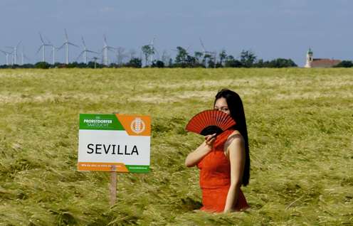 Frau im roten Kleid mit Fächer im Getreidefeld neben Schild „Probstdorfer Saatzucht Sevilla", Windräder im Hintergrund.