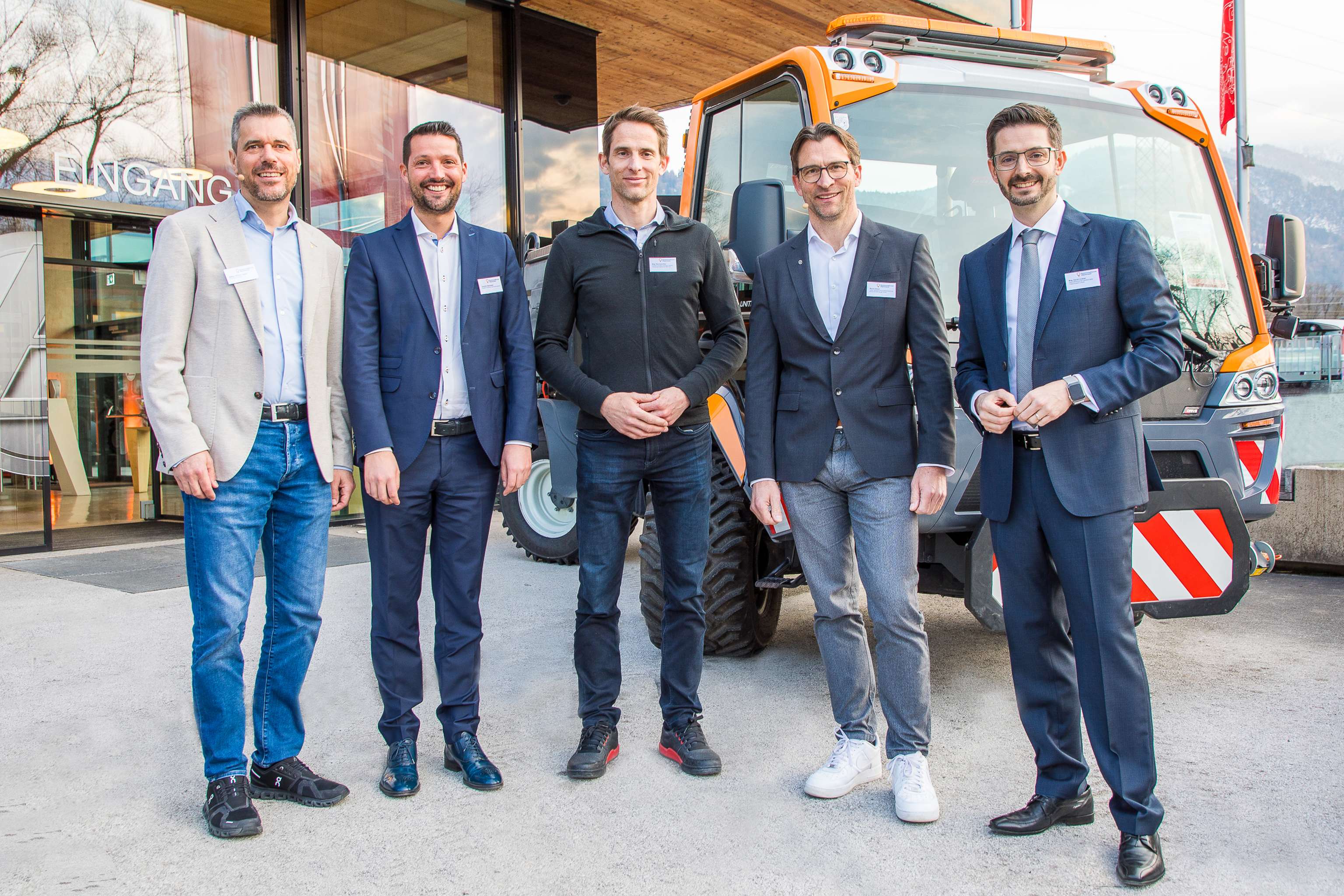 Fünf Männer in Geschäftsanzügen und Freizeitkleidung stehen vor einem gelben Fahrzeug, lächeln und posieren.