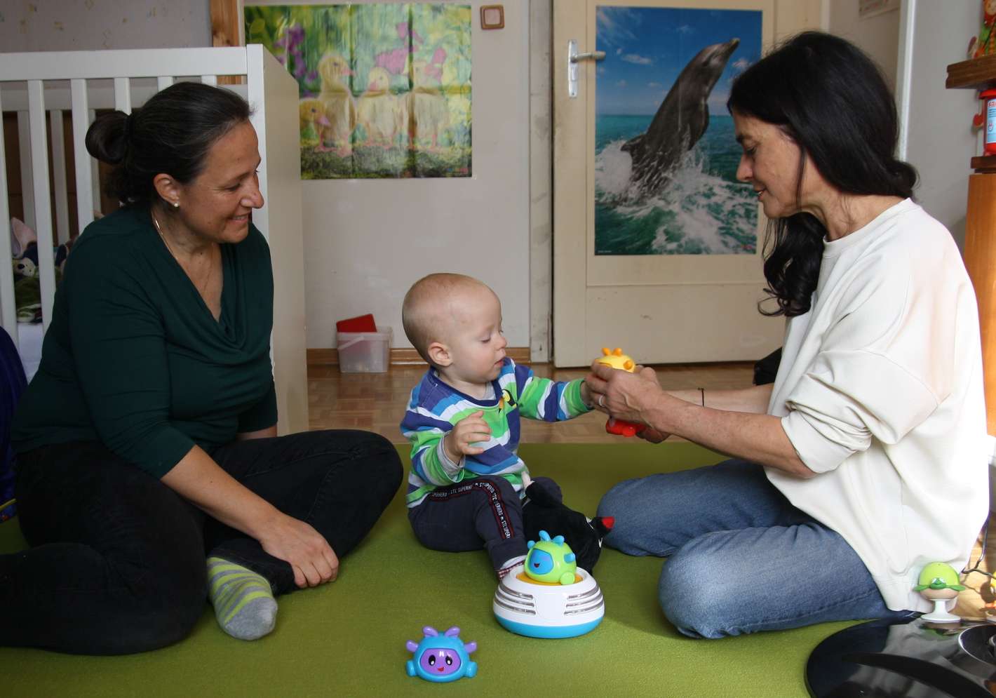 Zwei Erwachsene und ein Baby spielen auf dem Boden mit buntem Spielzeug in einem Kinderzimmer. Ein Poster mit einem Delfin hängt an der Tür.