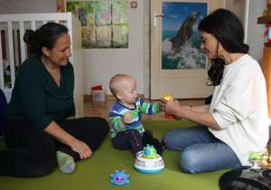 Zwei Erwachsene und ein Baby spielen auf dem Boden mit buntem Spielzeug in einem Kinderzimmer. Ein Poster mit einem Delfin hängt an der Tür.