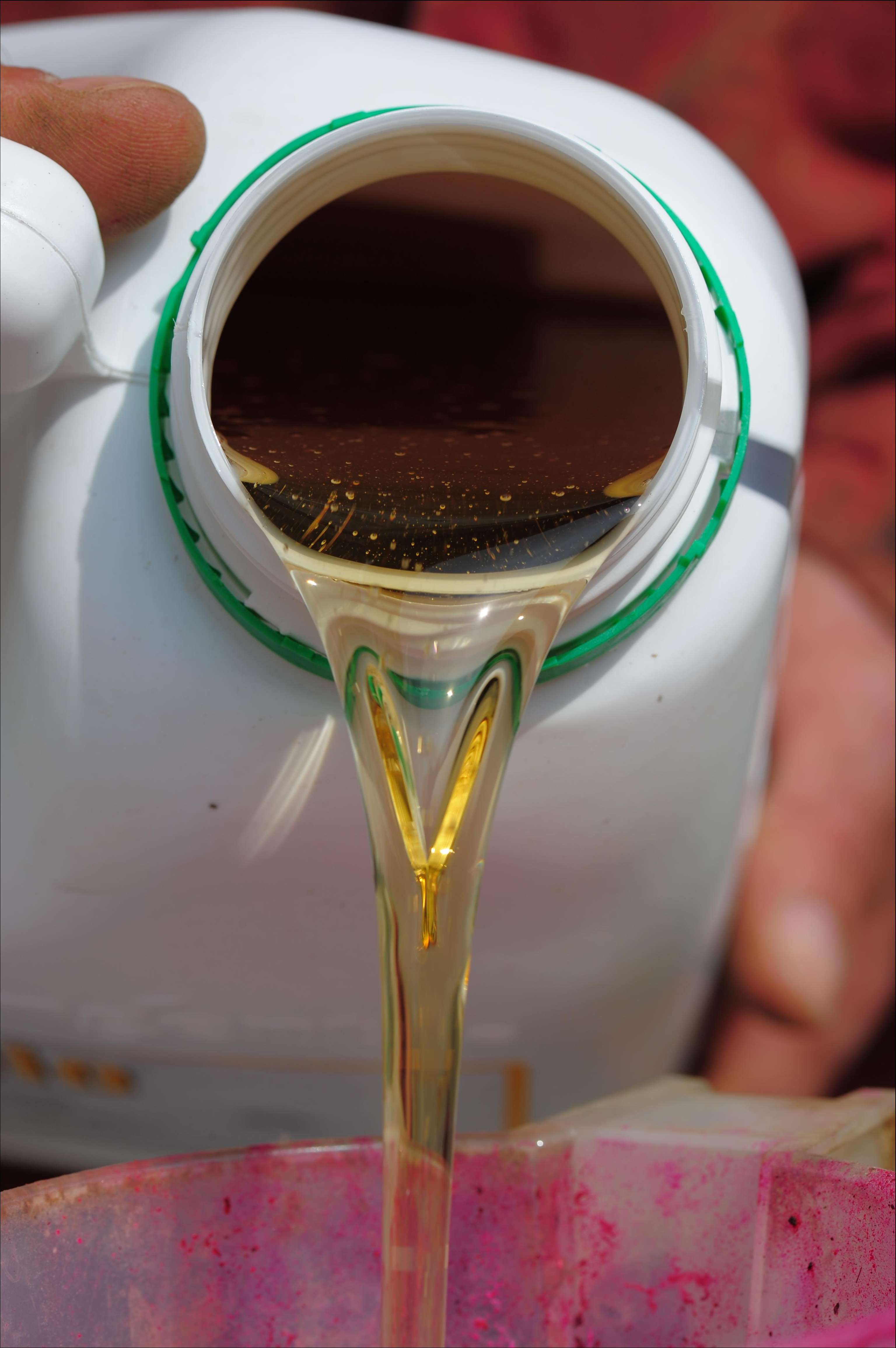 Close-up of a hand pouring golden liquid from a white container into a pinkish basin, with the liquid forming a smooth stream.