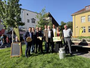 Gruppe von Personen bei einem Bio-Bauernmarkt im Freien, mit Käse und Bio-Zertifikat, Milchkanne und Tafel mit Aufschrift „Bio, regional und sicher".