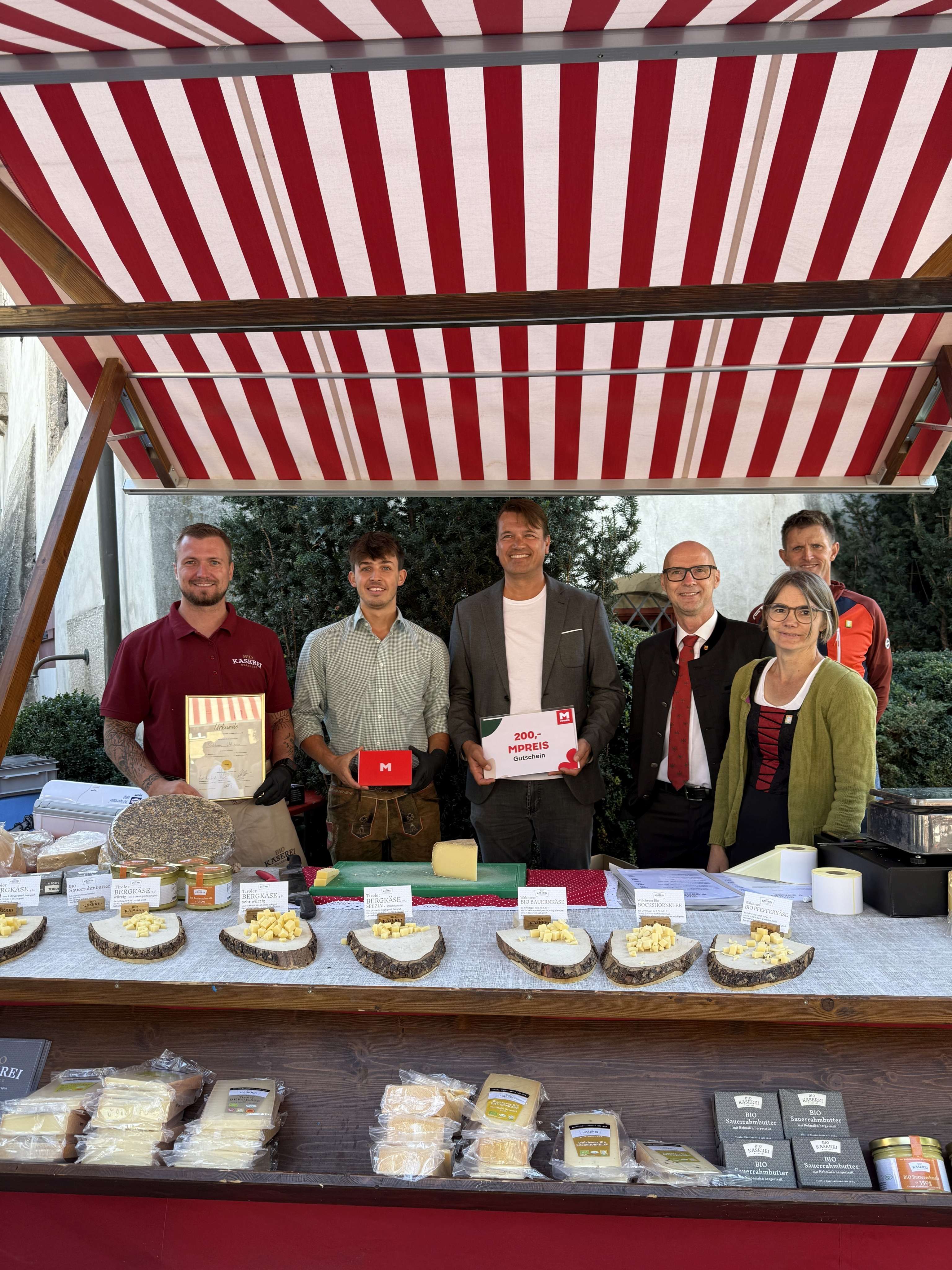 Fünf Personen an einem Käsestand mit rot-weißem Sonnendach, einer hält einen MPreis-Gutschein über 200 Euro, Bergkäse ausgestellt.