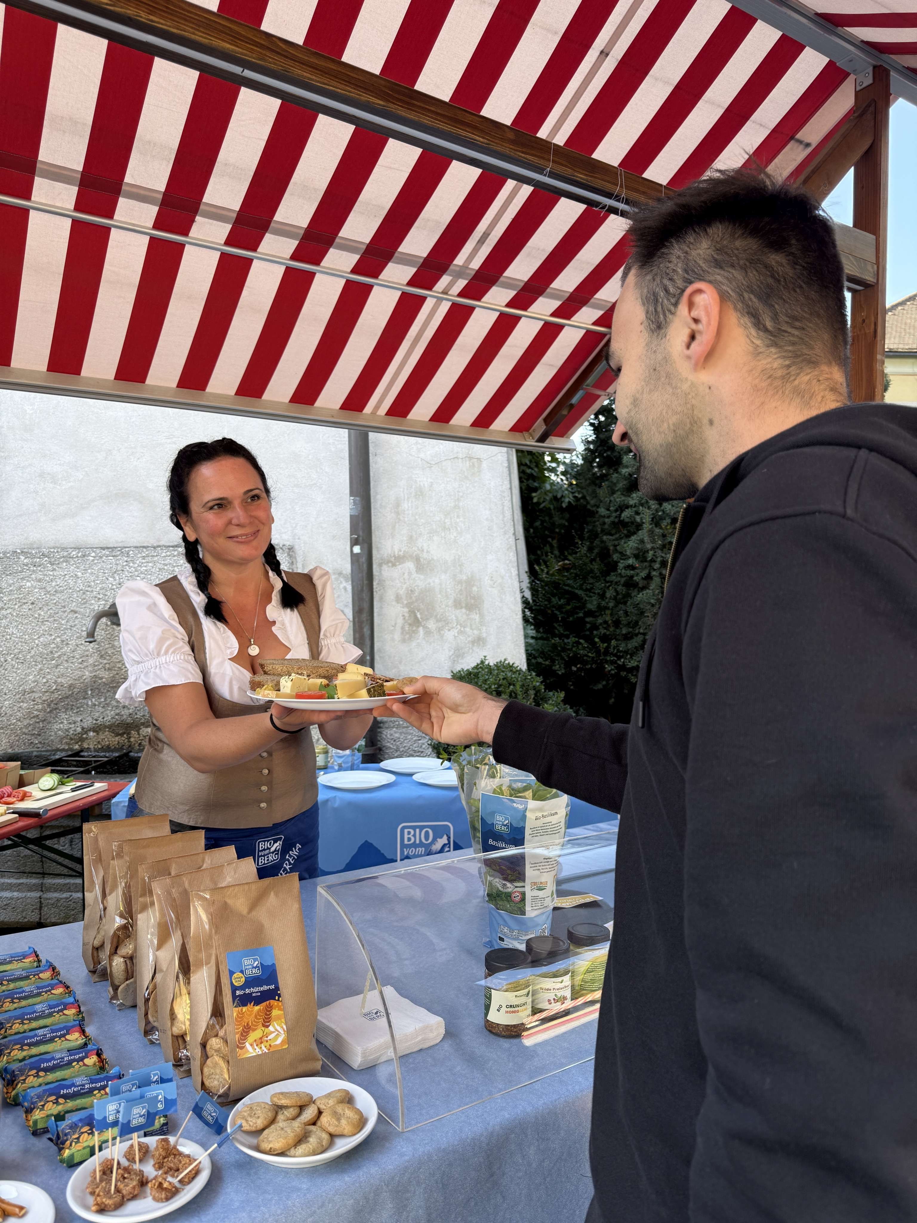 Frau in Tracht reicht einem Mann einen Teller mit Bio-Kostproben an einem Marktstand mit Bio vom Berg Produkten.