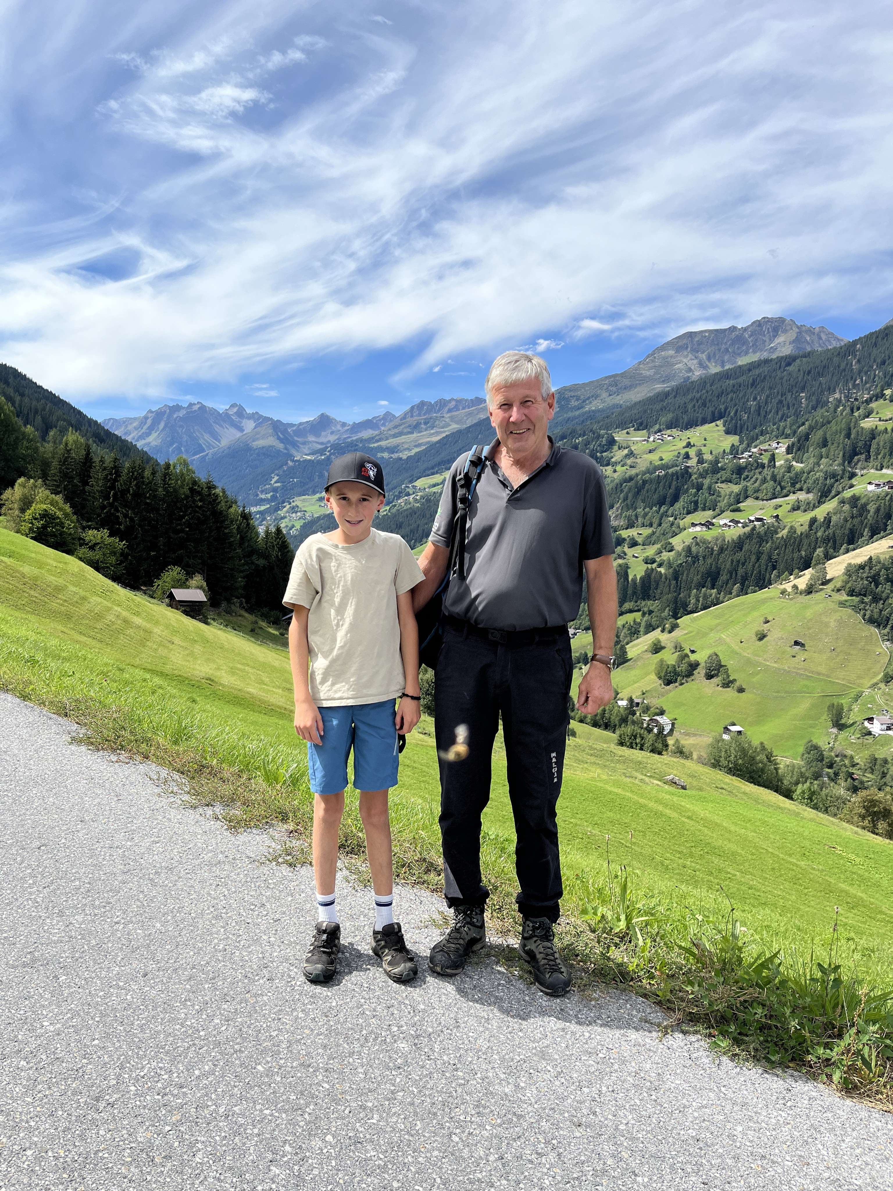 Ein älterer Mann und ein Junge stehen auf einem Bergweg in den Alpen, umgeben von grünen Wiesen und Berggipfeln unter blauem Himmel.