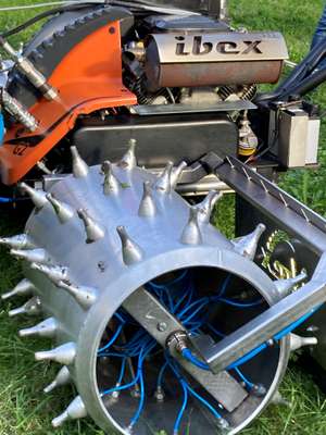 Maschine mit Metallzylinder, Stacheln und blauen Drähten auf Rasen. Aufschrift "ibex" sichtbar.