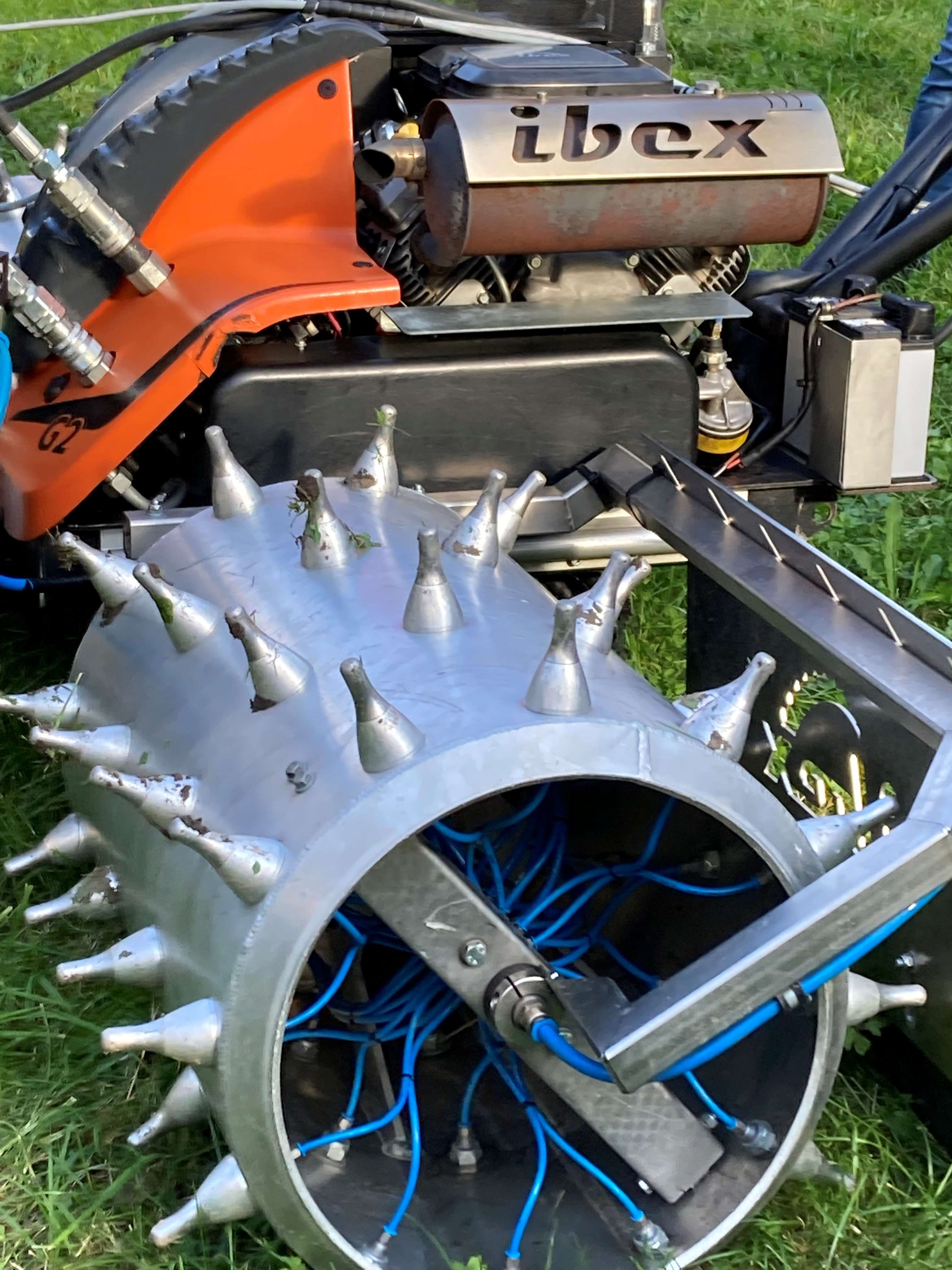 Maschine mit Metallzylinder, Stacheln und blauen Drähten auf Rasen. Aufschrift "ibex" sichtbar.