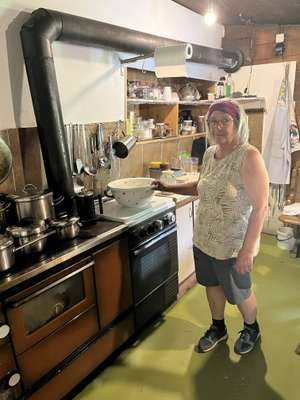 Ältere Frau mit Brille steht in rustikaler Küche neben einem alten Holzherd mit schwarzem Abzugsrohr und Kochgeschirr.