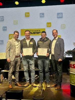 Four men stand on a stage holding certificates, smiling, with a large screen displaying logos in the background.