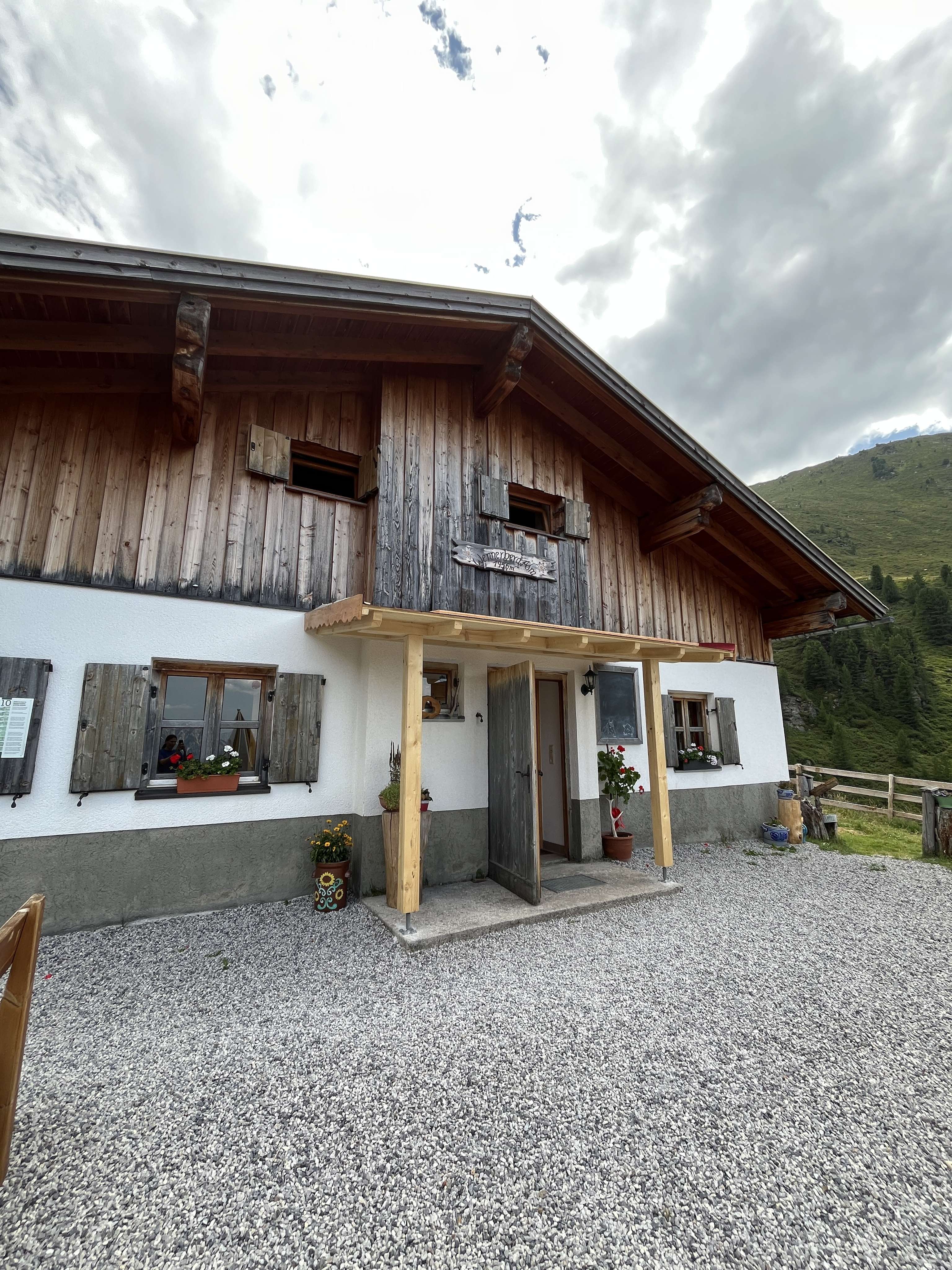 Traditionelle Almhütte mit Holzfassade, Fensterläden und Blumenschmuck in alpiner Berglandschaft unter bewölktem Himmel.