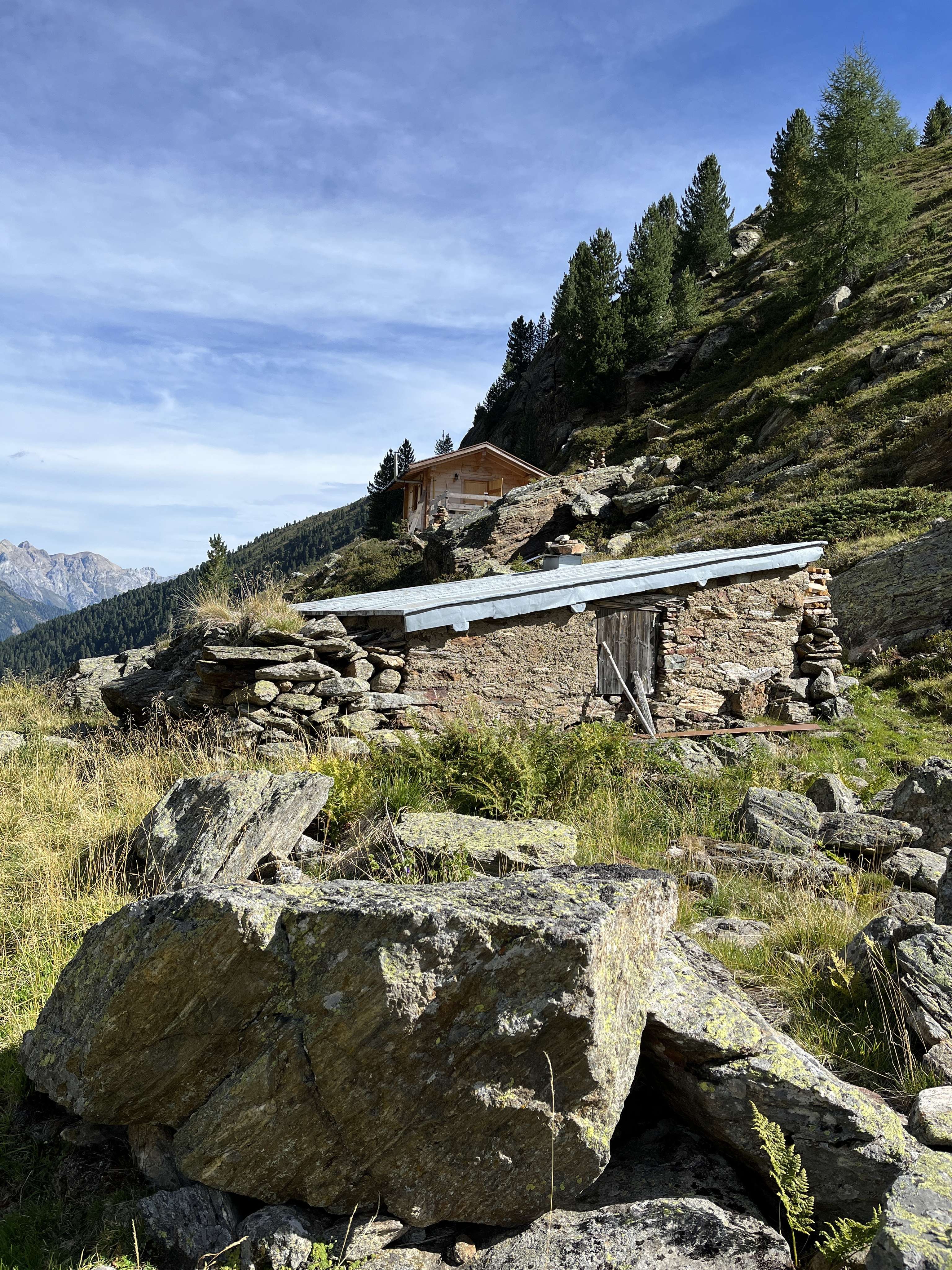 Alte Almhütte aus Naturstein mit Metalldach in alpiner Berglandschaft, umgeben von Felsen, Nadelbäumen und blauem Himmel.